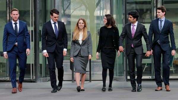 6 business persons walk towards the lens lens. The 2 business women are at the center of the group. 