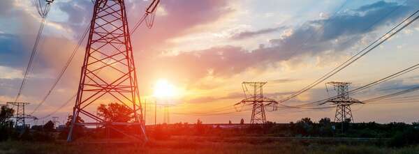 the sun shines between two electrics pylons. The sky is blue. Some big clouds are black. Clouds near the sun are orange.